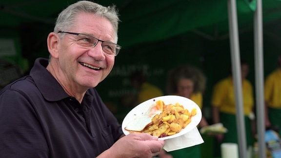 Ein älterer Mann mit Brille präsentiert auf einem Pappteller ein Kartoffelgericht. 