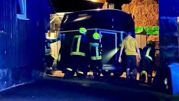 Feuerwehrmänner schieben bei Dunkelheit einen Tiertransport-Anhänger in einen Stall.