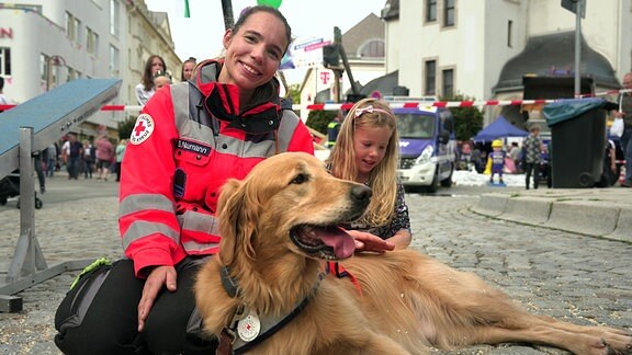 Eine Frau mit roter Rettungsweste sitzt mit einem Hund und ihrer Tochter auf der Straße. 