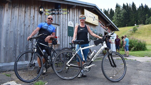 Vor einem Bistro 'Am Rölzhang' stehen ein Radfahrer und eine Radfahrerin in sportlicher Kleidung und Sonnenbrillen. Sie blicken in die Kamera. 