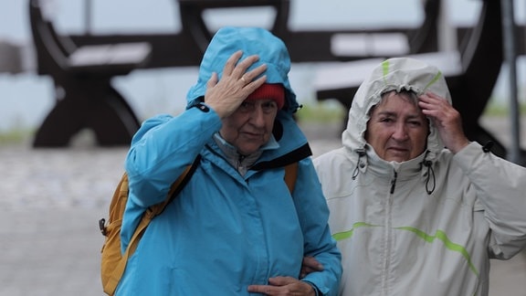 Zwei Wanderinnen gehen auf einer Bergkuppe durch Sturm. Sie haben sich ihre Regenkapuzen fest in die Gesichter gezogen und versuchen, die Kapuzen festzuhalten. 