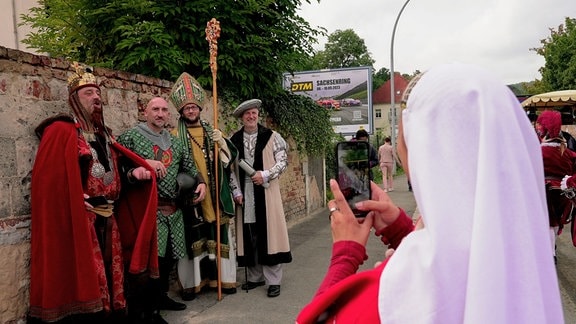 Vier in historischen Kostümen gekleidete Menschen stehen an einer Mauer und lassen sich von einer anderen Darstellerin mit dem Handy fotografieren.