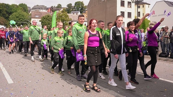 Menschen in lilafarbenen Sportsachen laufen als Gruppe auf der Straße und winken. Es sind Fans des 1. FC Erzgebirge Aue.