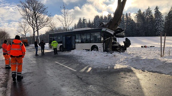 Ein Bus hat sich bei einem Unfall auf winterlicher Straße in einen Baum gebohrt, daneben stehen Männer mit orangener Kleidung. 