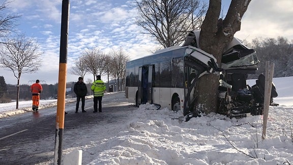 Ein Bus hat sich bei einem Unfall auf winterlicher Straße in einen Baum gebohrt, daneben stehen Männer mit orangener Kleidung. 