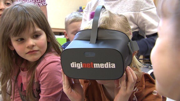 Ein kleines Mädchen mit VR-Brille wird von ihrer kleinen NAchbarin neugierig beobachtet