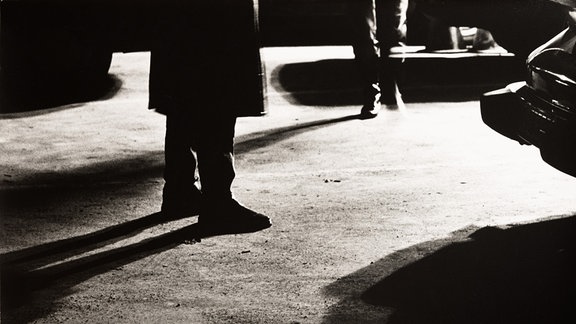 Fotografie von May Voigt: Angstnacht, 1989 - das schwarz-weiße Foto zeigt den Boden einer Garage, im Vordergrund die Beine eines Mannes, weiter hinten die Beine einer Frau