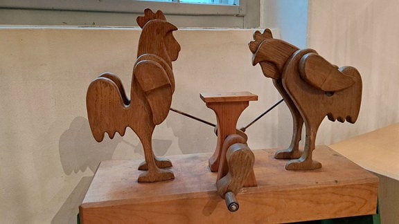 Ein Holzspielzeug: zwei Hühner, durch eine Kurbel angetrieben, picken auf einem Tisch. 
