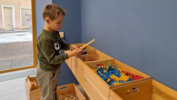 Ein Junge im Grundschulalter steht vor drei großen Holzkisten und greift in eine hinein, die voller Holzspielzeug und Bauteilen ist.