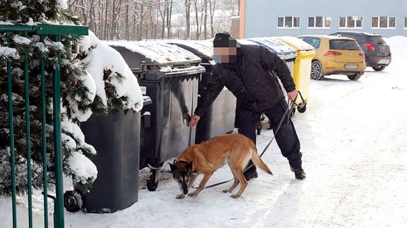 Ein Polizist steht mit einem Spürhund vor Mülltonnen.