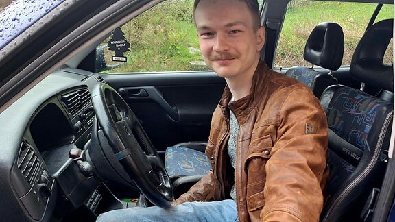 Ein junger Mann in Jeans und Lederjacke steigt in sein Auto und schaut freundlich in die Kamera.