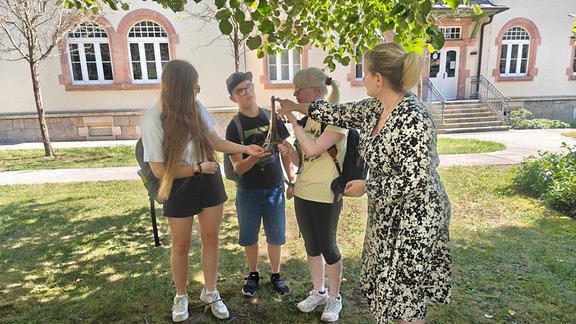 Zwei Schülerinnen, ein Schüler und eine Lehrerin stehen zusammen auf einer Wiese. Im Hintergrund ist ein Gebäude zu sehen. Alle vier Personen halten bzw. berühren ein Tastmodell eines Eifelturms. 