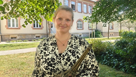 Eine Frau im Weiß-schwarz gemusterten Kleid steht unter Bäumen auf einer Wiese vor einem Gebäude. Sie hält ein Tastmodell vom Eifelturm in der Hand und lächelt in die Kamera.