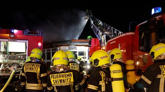 Feuerwehrleute neben einem Drehleiterfahrzeug, von dessen Spitze Kameraden ein brennendes Dach löschen.