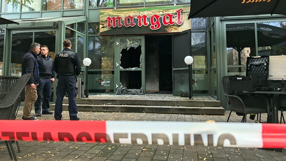 Hinter einem Absperrband der Polizei ist die zerstörte Tür eines Restaurants zu sehen