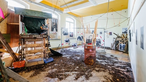 Fotografien hängen an einem Bauzaun, Dachlatten stecken in einem Fass, Blätter von Bäumen liegen auf dem Boden.