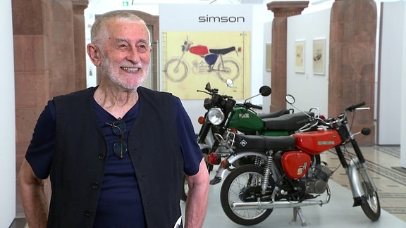 Clauss Dietel, ein Mann mit grauen Haaren und Bart steht lachend in einer Ausstellung vor Motorrädern