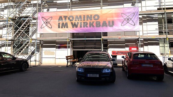 Autos stehen vor einem eingerüsteten Rohbau, auf einem lila-orangenen Banner steht "Atomino im Wirkbau":