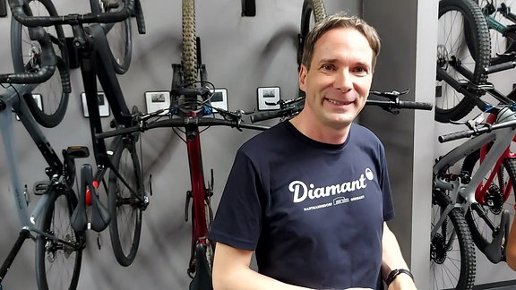 Ein Mann Mitte 40 steht in einem dunklen T-Shirt vor einer Wand, an der mehrere neue Fahrräder hängen. Es ist der Geschäftsführer des Fahrradherstellers Mirco Schmidt in Hartmannsdorf. 