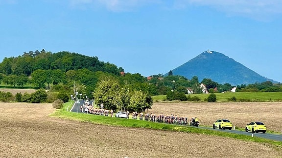Auf einer Landstraße fahren Radsportler und Begleitfahrzeuge eines Etappenrennens. Dahinter ein steiler Berg mit Burg.