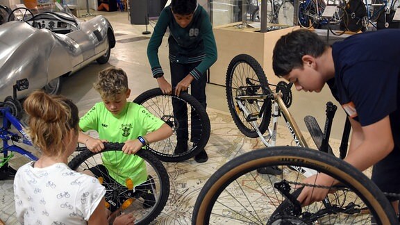 Drei Jungs mit demontierten Fahrrad-Rädern am Boden einer Ausstellungshalle.