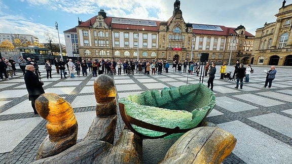 Blick über die Skulptur "Glasarche", ein Schiff und eine Hand auf Zuschauer, die im Halbkreis davorstehen. 