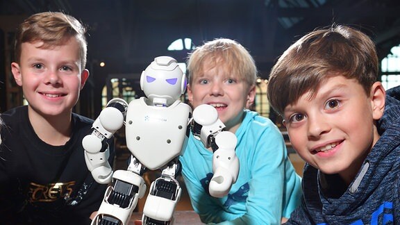 Drei Jungs an einem Tisch mit kleinem Roboter und Minifußball in einer alten Industriehalle 