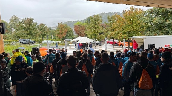 Eine große Gruppe steht im Regen unter einem Dach und schaut nach draußen. 