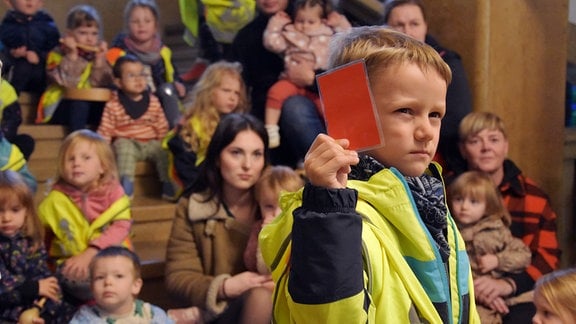 Ein Kindergartenkind zeigt eine rote Karte in die Kamera, dahinter sitzen andere Kinder