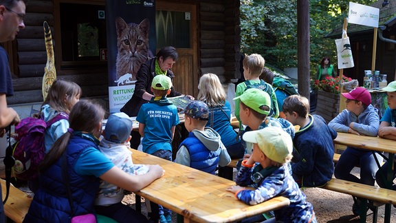 Eine ältere Frau zeigt einer Kindergruppe ein Bild. Die Kinder sitzen auf Bierbänken und schauen zu ihr.