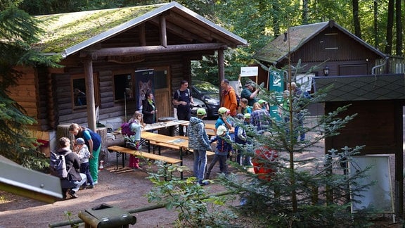 Vor einer Holzhütte tummelt sich eine Gruppe von Kindern. Ein paar Erwachsene stehen dazwischen.