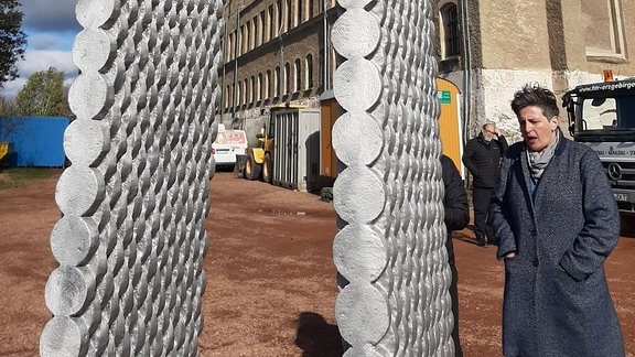 Die Skulptur ist ein glänzender Torbogen von etwa zwei Meter Höhe. Daran lehnt die Künstlerin in Winterjacke. Im Hintergrund sind die Mauern der Festung Hoheneck zu sehen. 