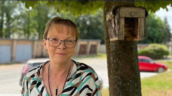 Eine Frau steht neben einem Baum uns schaut in die Kamera.