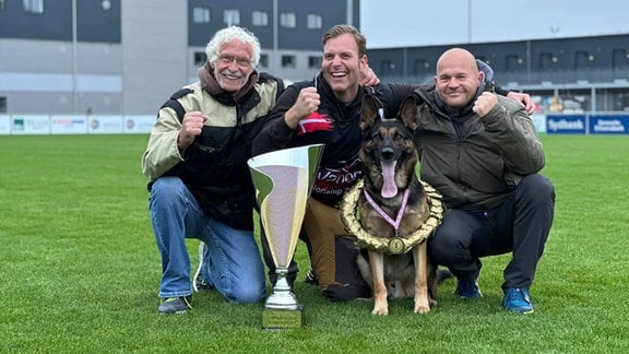 Drei Männer posieren mit einem Hund und einem Pokal.