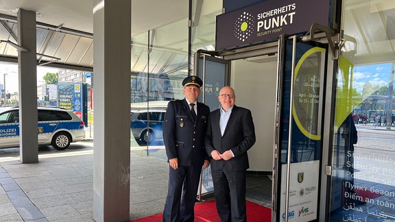 Der Chemnitzer Polizeipräsident und der Chemnitzer Oberbürgermeister stehen vor einer geöffneten Doppeltür auf einem roten Teppich und lächeln in die Kamera. Über der Eingangstür steht "Sicherheitspunkt"