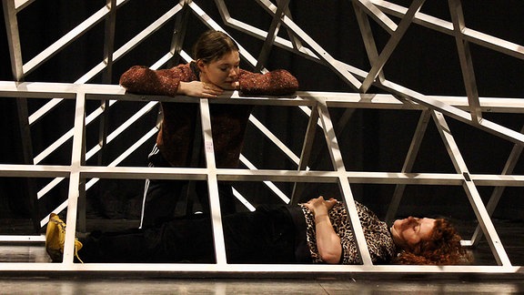 Szene aus einem Theaterstück: zwei Frauen stehen in einem Gerüst, das aussieht wie ein Haus und schauen sich an.