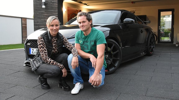 Ein Mann und eine Frau hocken vor einem schwarzen Sportwagen Audi TT RS vor einer Garage.