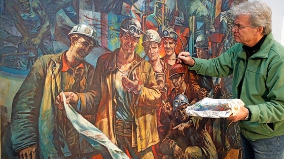 Der Berliner Maler Werner Petzold, ein älterer Mann mit grauen Haaren, Brille und grüner Jacke, restauriert mit Pinsel und Farbe das von ihm vor 40 Jahren geschaffene Gemälde "Brigade Rose" im Kunstarchiv der Wismut GmbH in Chemnitz.