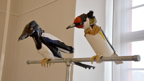 Zwei Vögel aus gesammelten Abfällen sitzen auf einer Stange in einem Ausstellungsraum. 