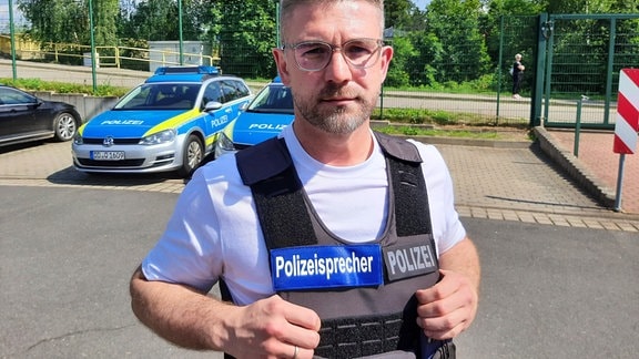 Der Sprecher der Polizeidirektion Chemnitz, Andrzej Rydzik, steht mit einer Polizeiweste vor zwei Polizeiautos.