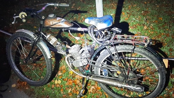  Ein Fahrrad, an dem nachträglich ein Mopedmotor angebaut wurde, lehnt an einem Zaun. 