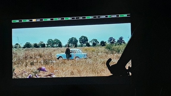 Blick auf eine Leinwand bei Nacht. Zu sehen ist ein blauer Trabbi im Feld. Im Vordergrund die tretenden Zuschauer auf Fahrrädern. 