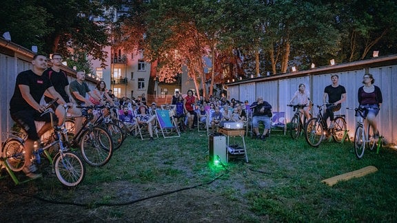 Fahrradkino im Garagenhof: Links und rechts sitzen Menschen auf Fahrrädern und strampeln. Dahinter weiteres Publikum auf Liegestühlen, in der Mitte der Beamer. Alle schauen in Richtung Kamera. 