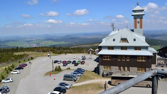 Blick von einem Berg in ein Tal bei blauem Himmel. Im Vordergrund steht ein Hotel, vor dem mehrere Autos parken.