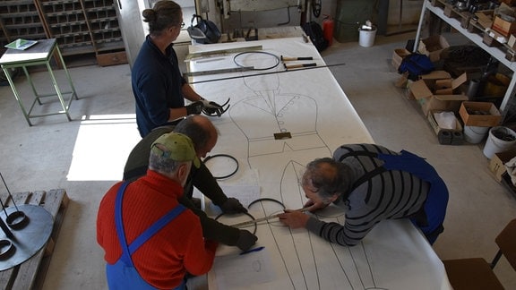 Vier Männer passen auf einem Tisch auf einer Zeichnung eines Bergmanns im Maßstab 1:1 Metallteile an.