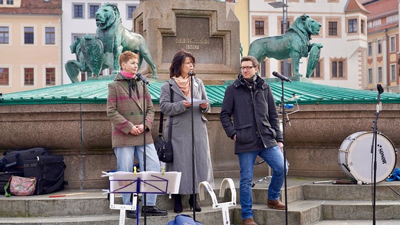 Drei Personen stehen bei einer Kundgebung hinter Mikrofonen.