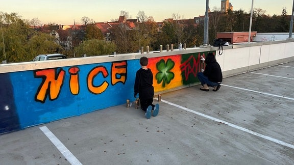 (Zwei Personen in schwarzer Kleidung sprühen an einer Wand ein Grafitti.)