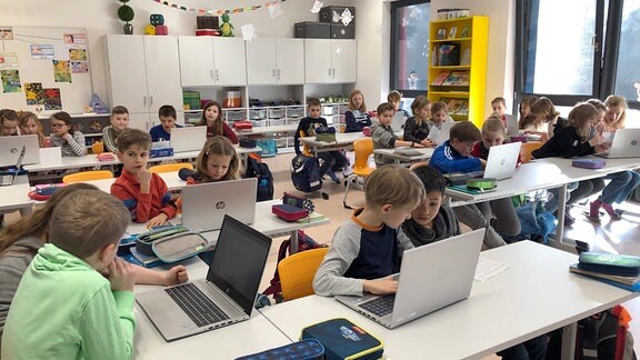 Kinder vor Laptops an der Schulbank sitzend. 