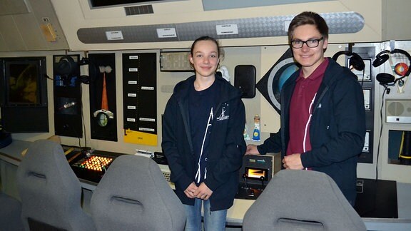 Ein Mädchen und ein Junge stehen vor einer Schalttafel,die das Cockpit eines Raumschiffes simuliert