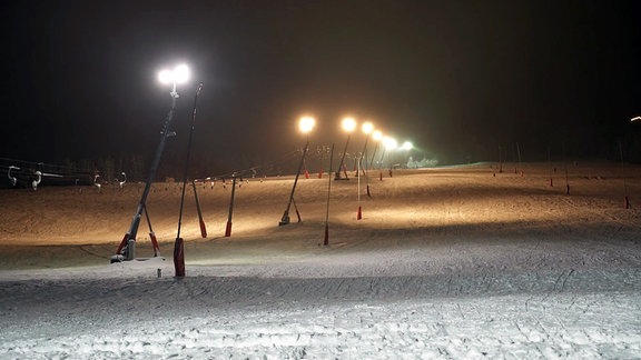 Ein menschenleerer, hell erleuchteter Skihang mit Schlepplicht bei Nacht.